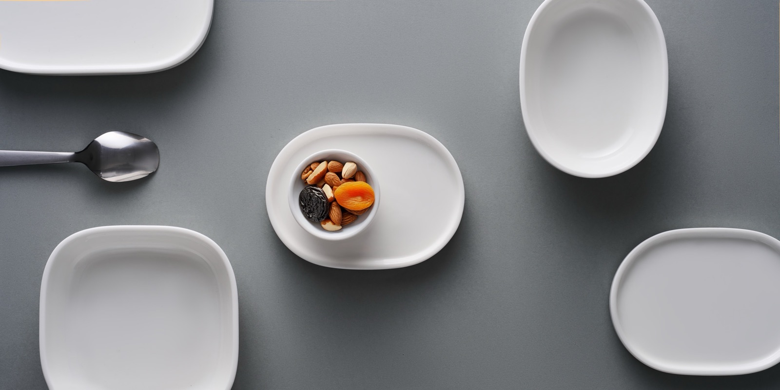 Flat-lay of the dinnerware system on a grey surface - square and oval plates, bowls, a snack dish, and spoon arranged with generous spacing.