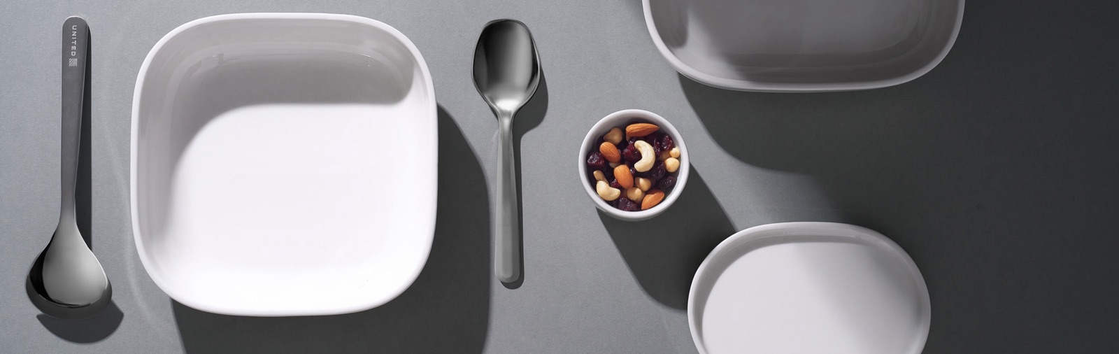 Close detail of the dining system on grey - a United-branded spoon, square bowl, and a small dish of dried fruit and nuts.