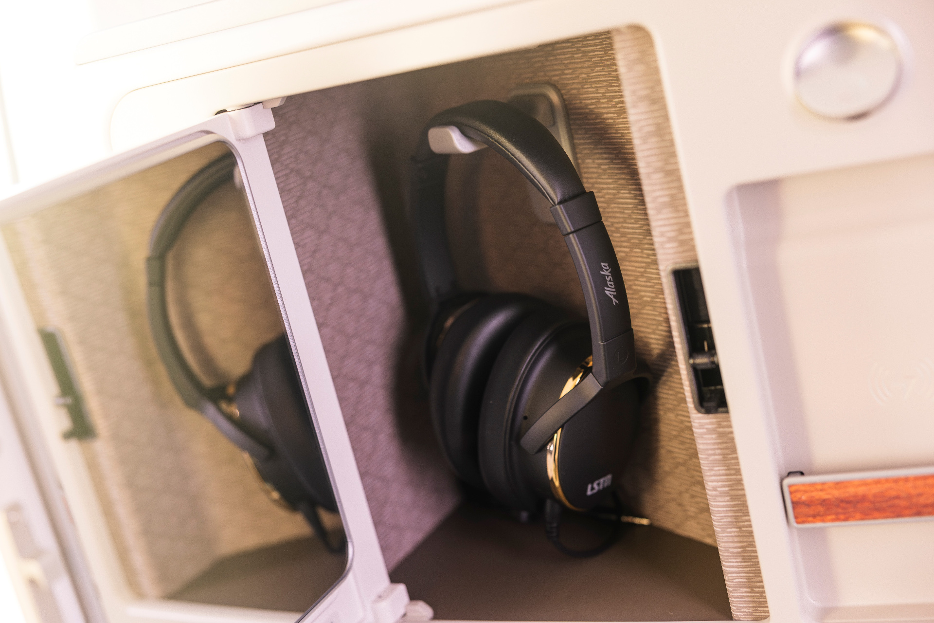 The headphones stowed in an Alaska Airlines Business Class seat compartment, the Alaska script visible on the headband against the cabin trim.