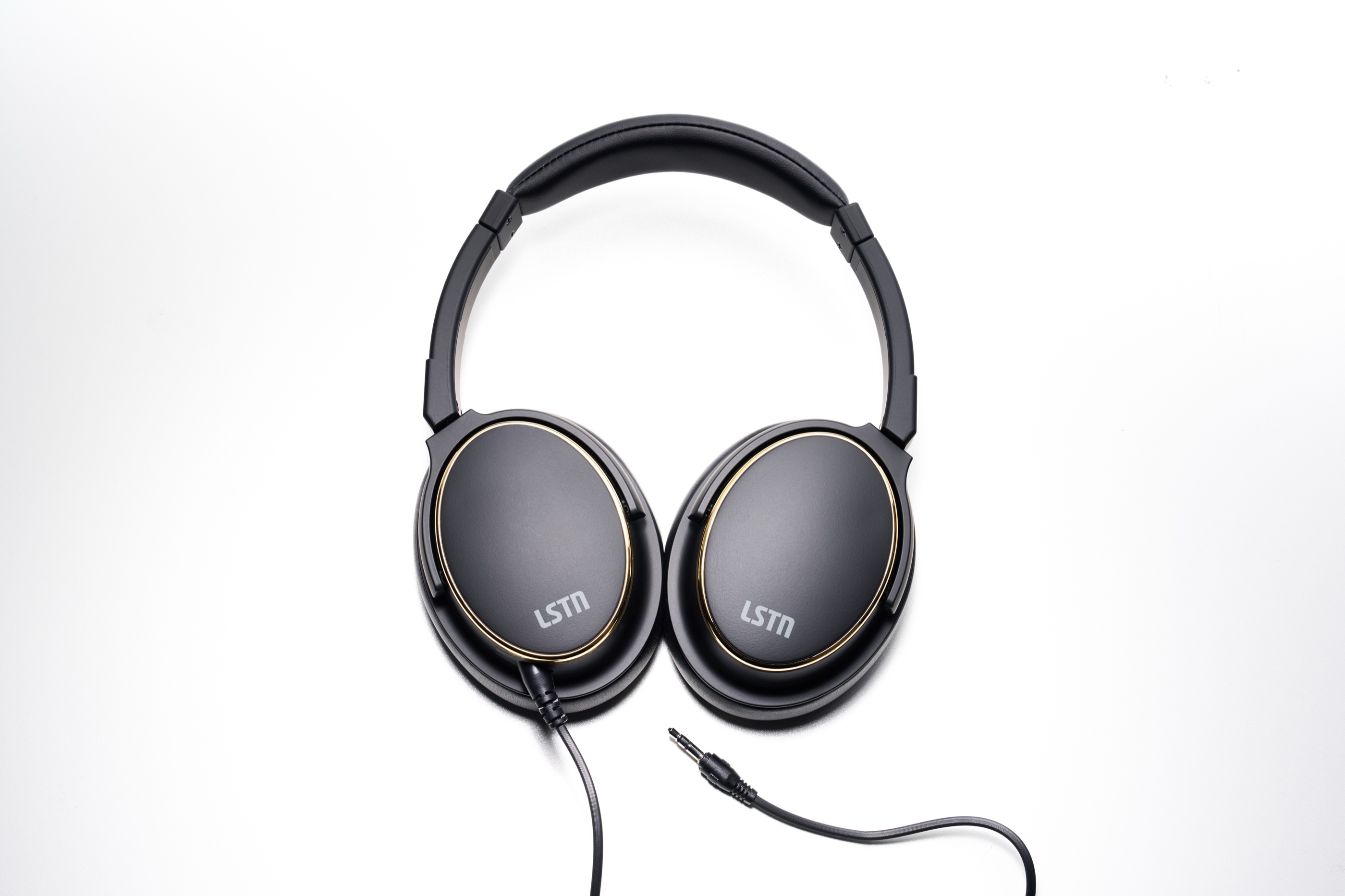 An overhead view of the headphones laid symmetrically on white - both matte black earcups ringed in brass with LSTN branding, the headband arcing above and the detachable 3.5 mm single-pin cable and connector below.