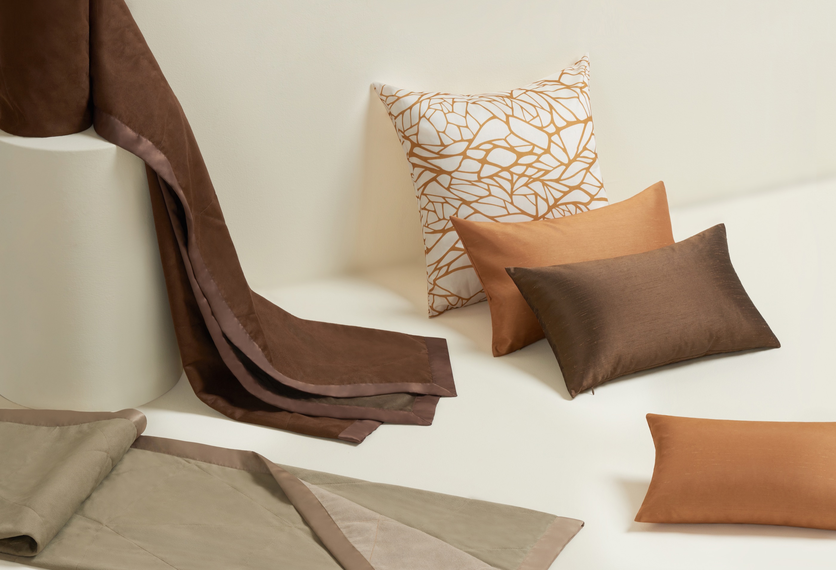 The Etihad bedding collection arranged as an editorial flat-lay - a draped brown throw, the bronze vein-pattern cushion, copper and brown pillows, and taupe linens.