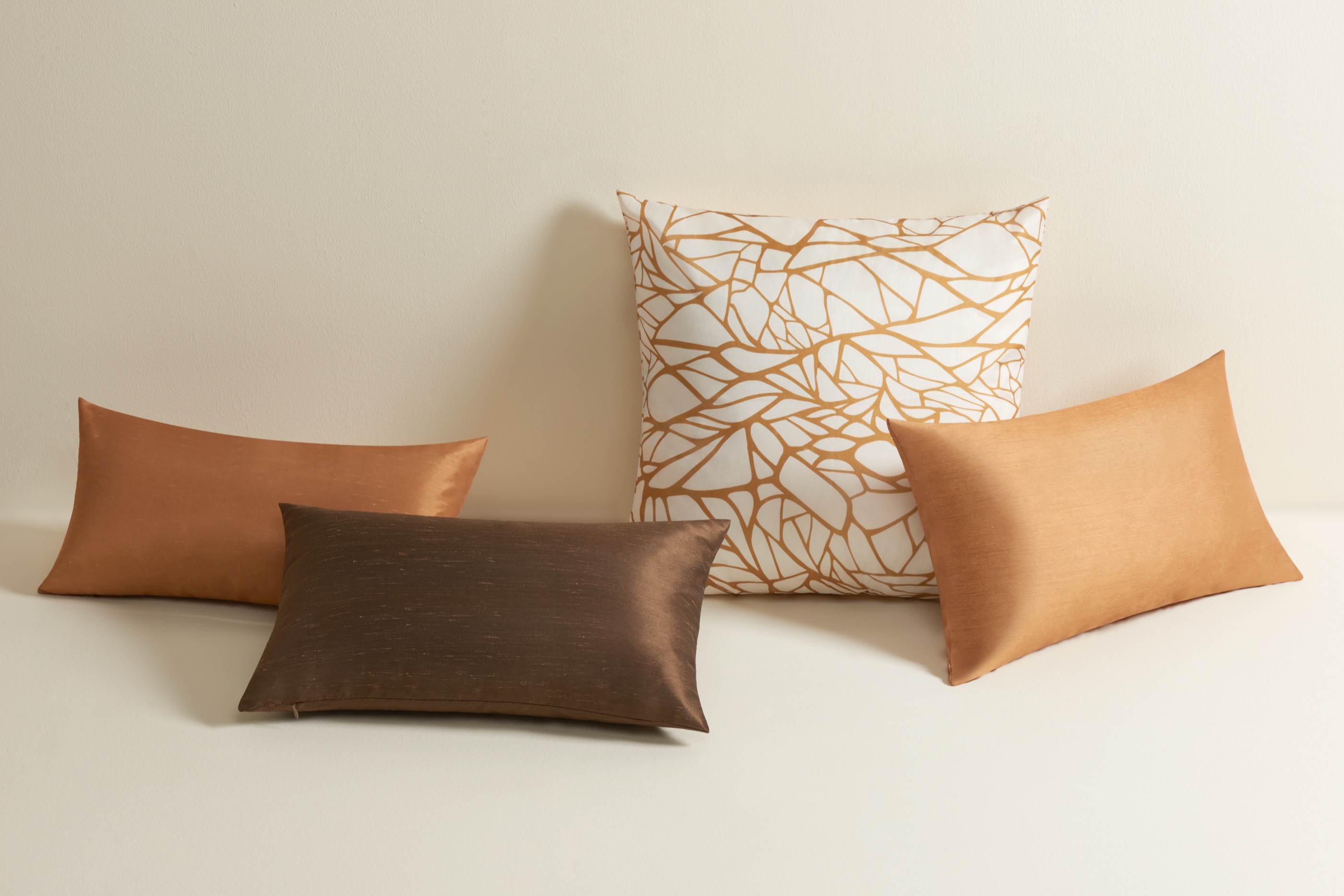 Four Etihad cushions - a white pillow printed with an interlacing bronze vein pattern, flanked by copper, brown, and tan solid cushions.