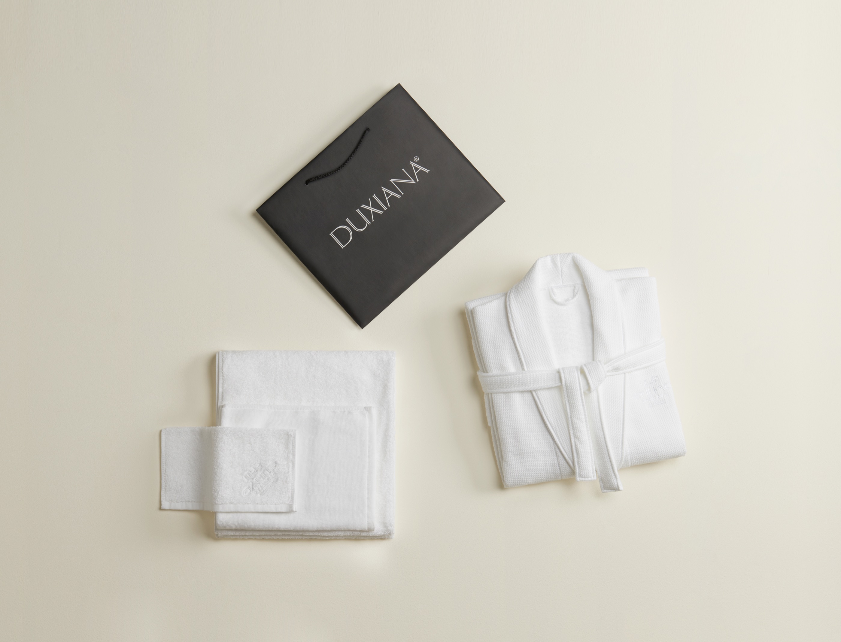 DUXIANA soft goods for Etihad - a black DUXIANA gift bag, a folded white waffle robe, and stacked white towels arranged on a cream ground.