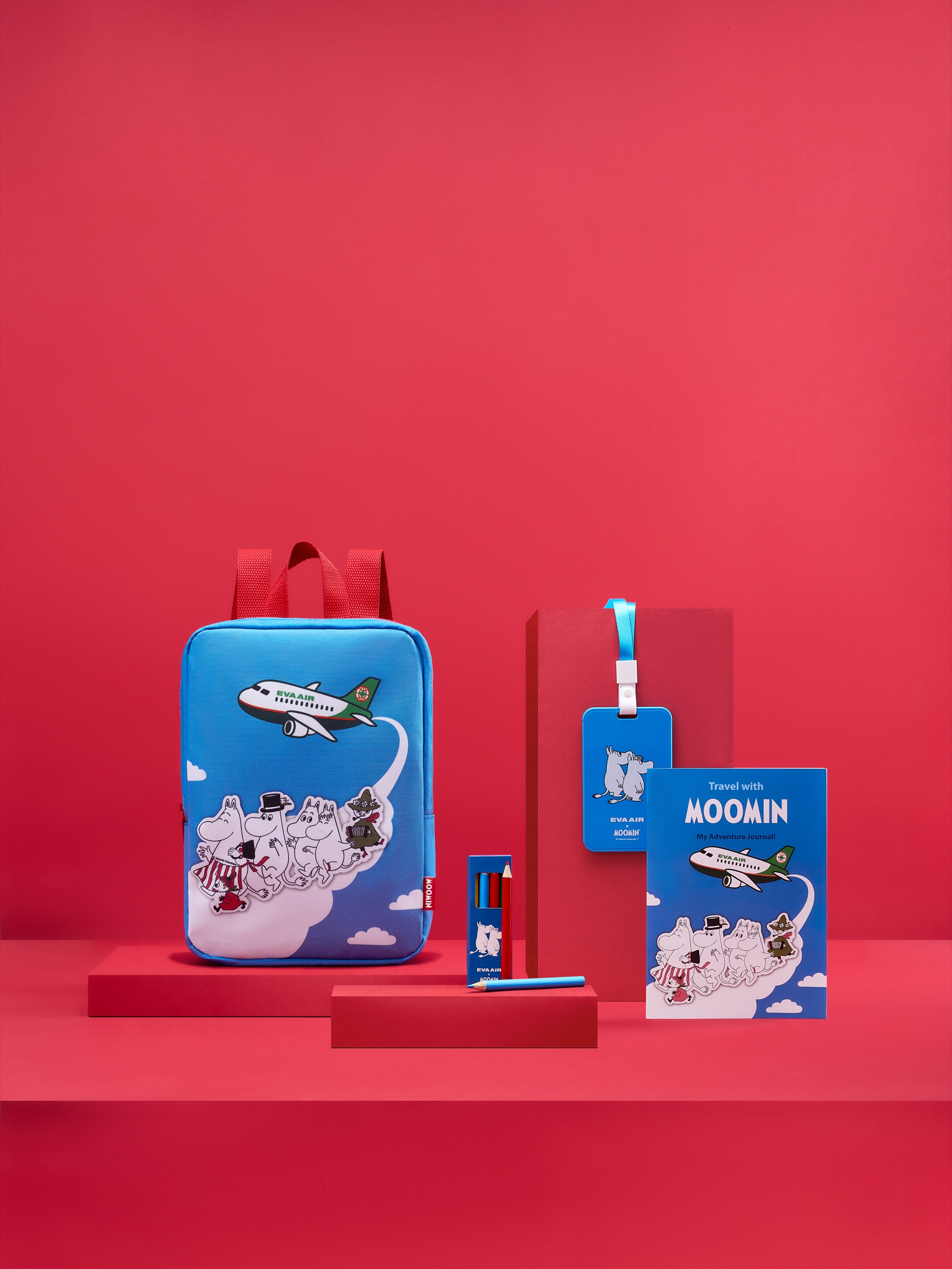 The full EVA Air and Moomin kit laid out on a red set - a blue backpack printed with an EVA Air aircraft and Moomin characters, a matching luggage tag, colour pencils, and a &lsquo;Travel with Moomin&rsquo; activity book.