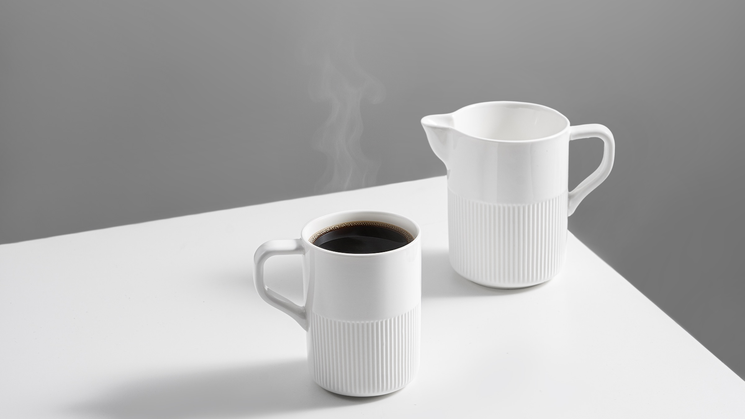 The fluted mug and coffee pot on a white surface, steam rising from freshly poured coffee.