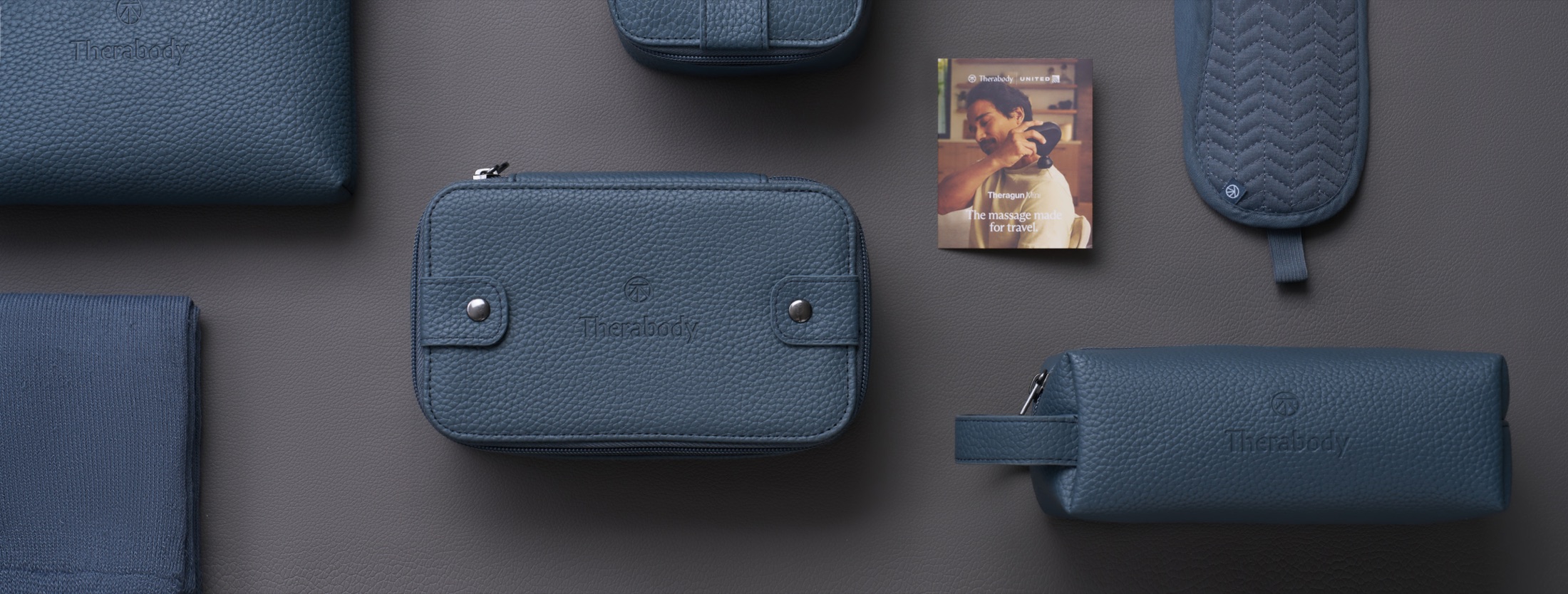 Wide overhead flat-lay of the navy Polaris kit on a dark surface - pebbled leatherette pouches, a quilted eye mask, socks, and the Theragun Mini insert card.