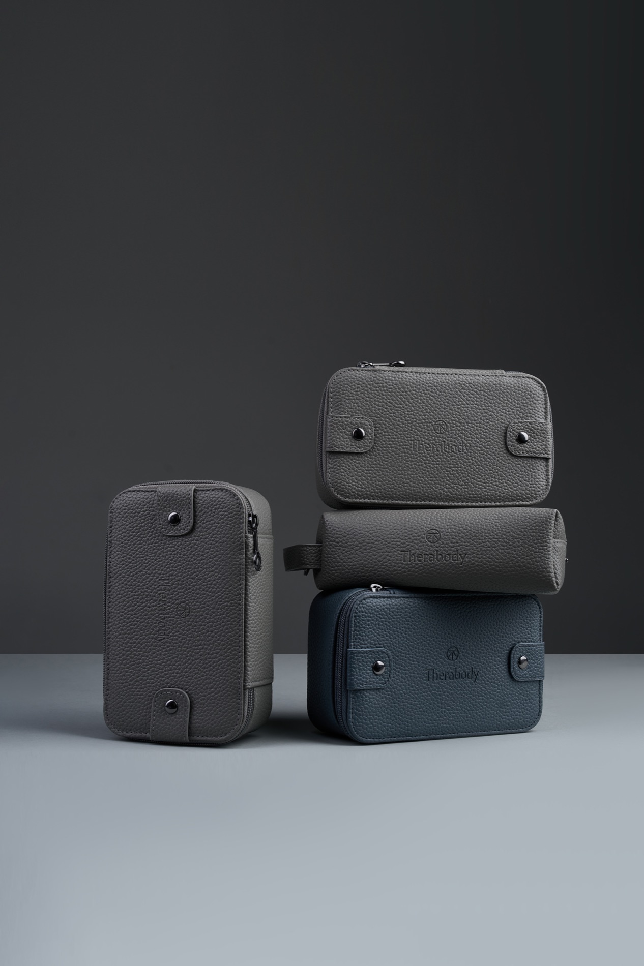 Three pebbled recycled-leatherette Therabody bags in grey and navy stacked against a dark backdrop, showing the paired collectible structures of the final rotation.