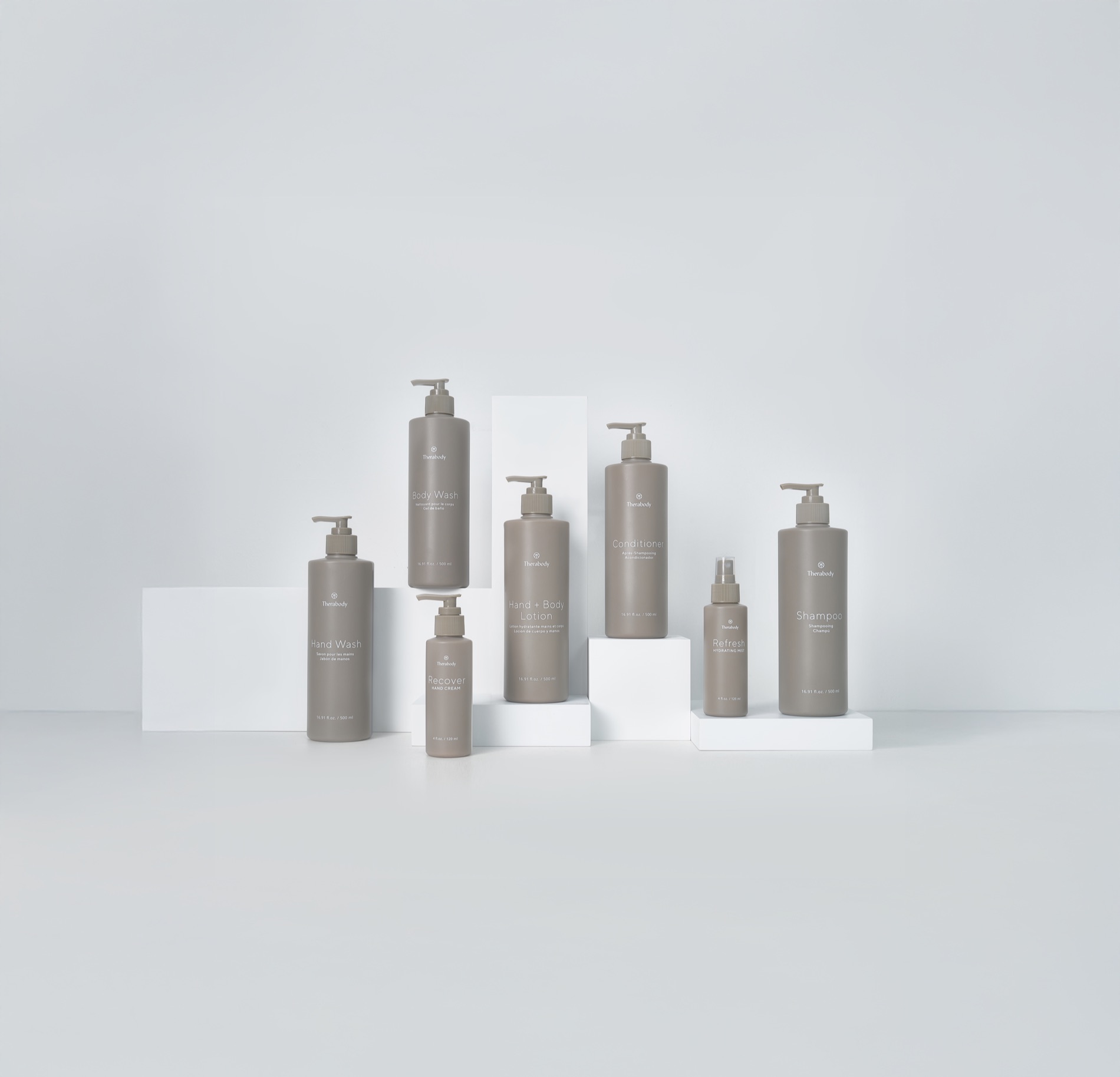The full-size Therabody lavatory amenity range - hand wash, body wash, shampoo, conditioner, hand and body lotion, and refreshing mist - arranged on stepped white plinths.