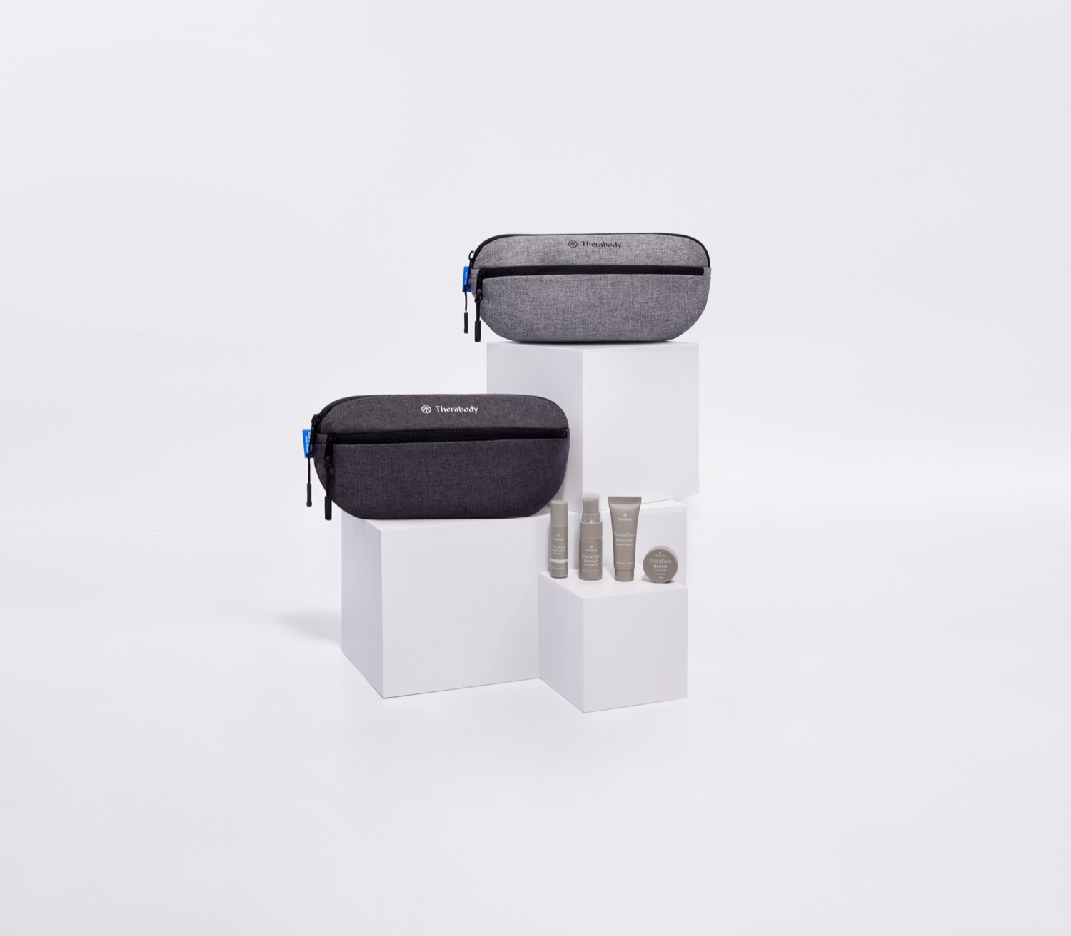 Two heather-grey RPET Therabody wash bags on white pedestals alongside the skincare minis, showing the recycled material strategy.
